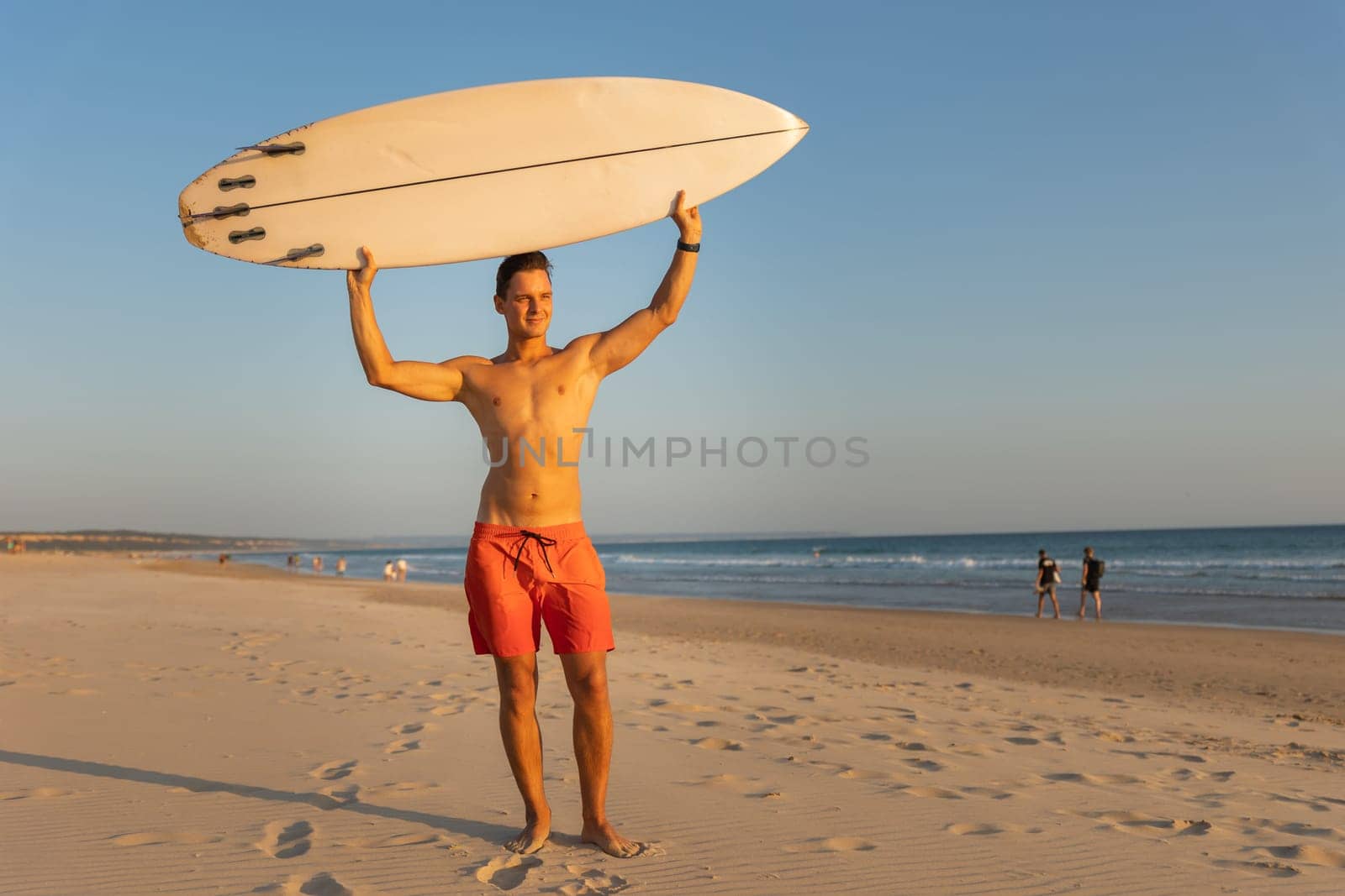 An attractive man with nice athletic body standing on the shore holding a surfboard above his head. Mid shot