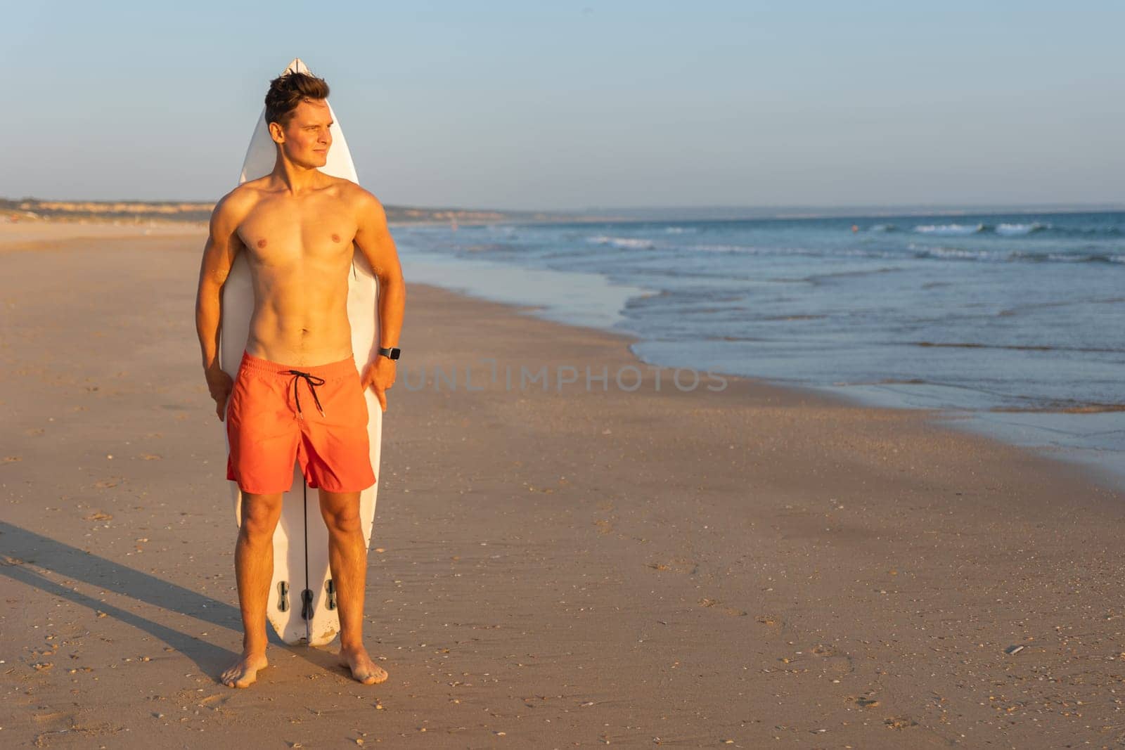 An attractive man with nice athletic body standing on the shore with a surfboard behind his back. Mid shot
