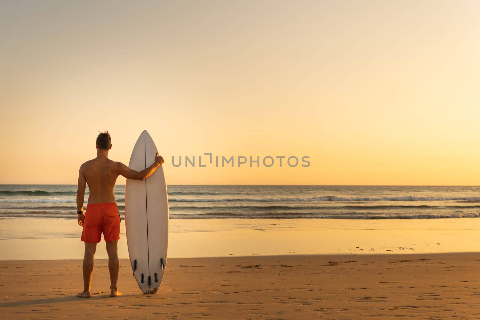 A man standing on the shore holding a surfboard - view from the back. Mid shot