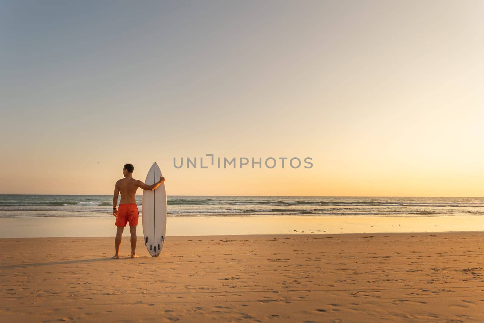 A man with athletic body standing on the shore holding a surfboard at early sunset - view from the back. Mid shot