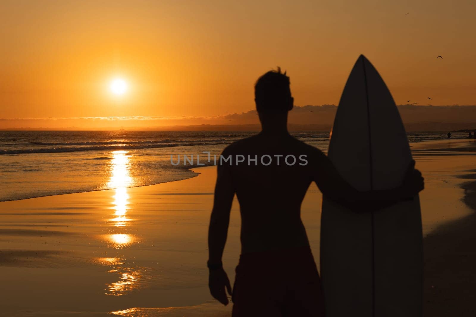 Black silhouette of an athletic man standing on the shore holding a surfboard and looking at sunset. Mid shot