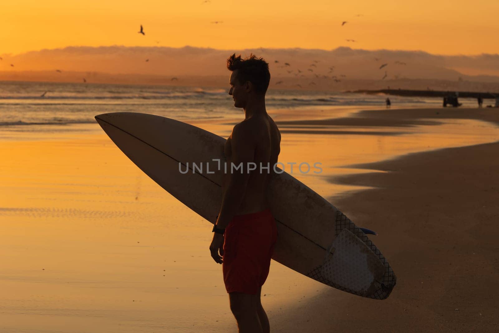 Black silhouette of an athletic man standing on the shore holding a surfboard at sunset. Mid shot