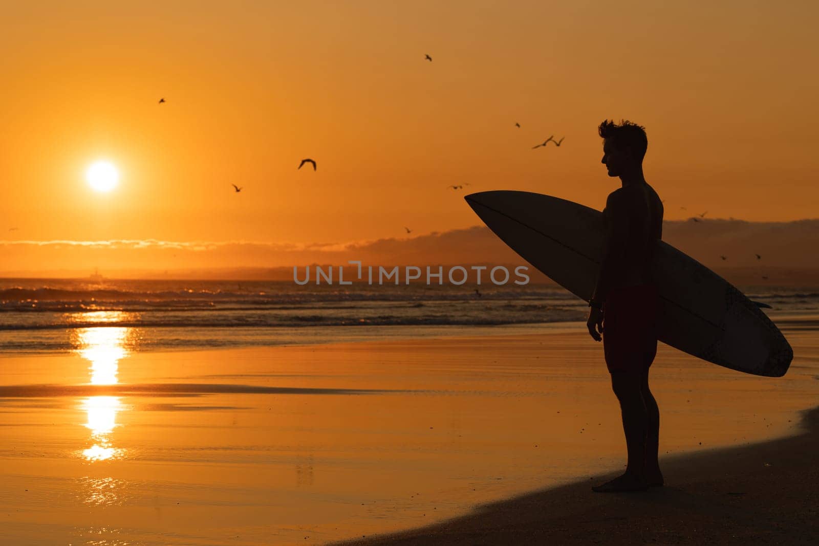 Black silhouette of an athletic man standing on the shore holding a surfboard at orange sunset. Mid shot
