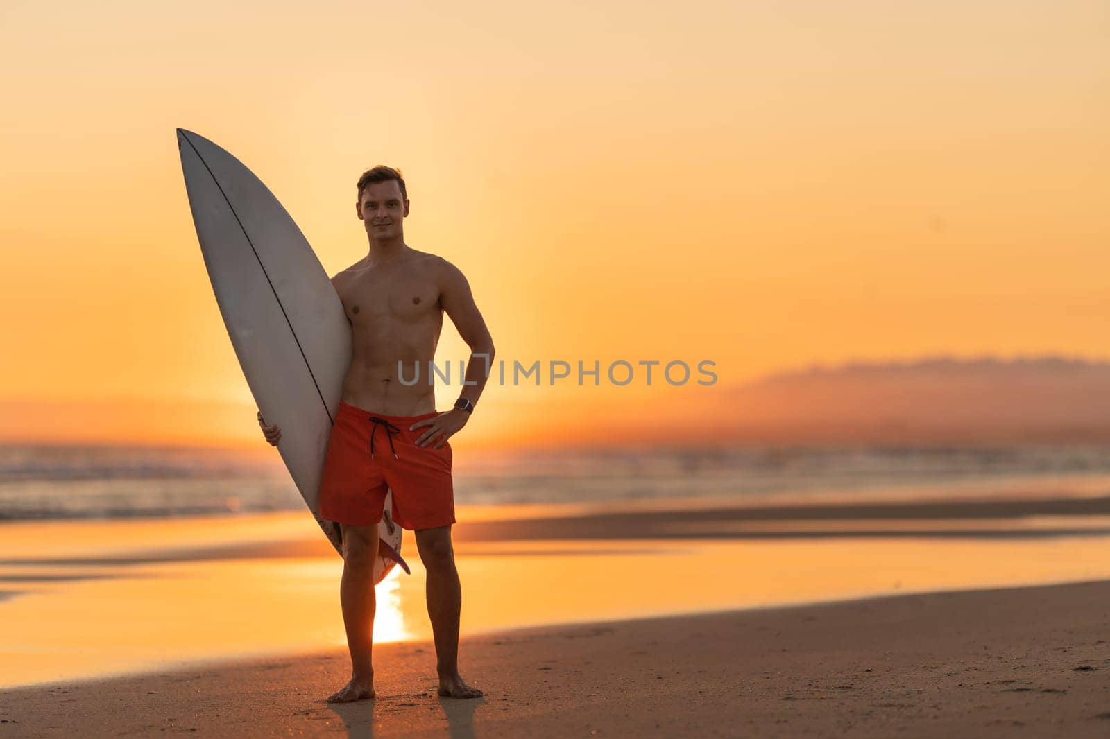 An attractive man standing on the shore holding a surfboard at orange sunset by Studia72