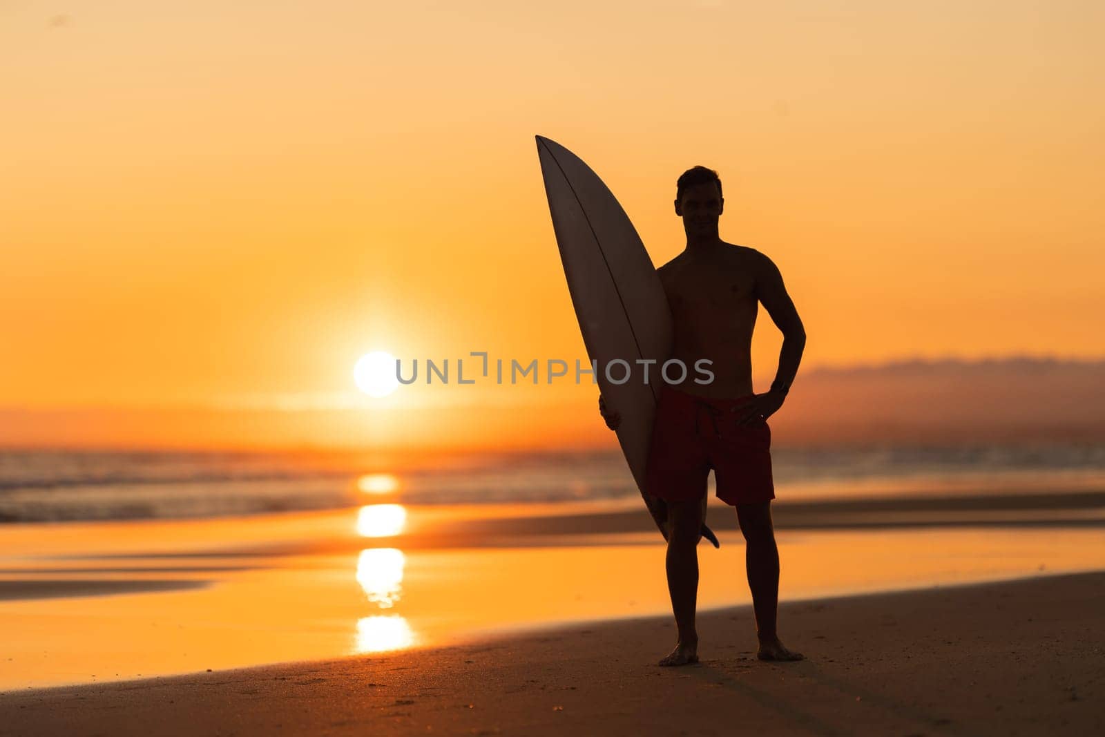 Black silhouette of an attractive man standing on the shore holding a surfboard at orange sunset. Mid shot