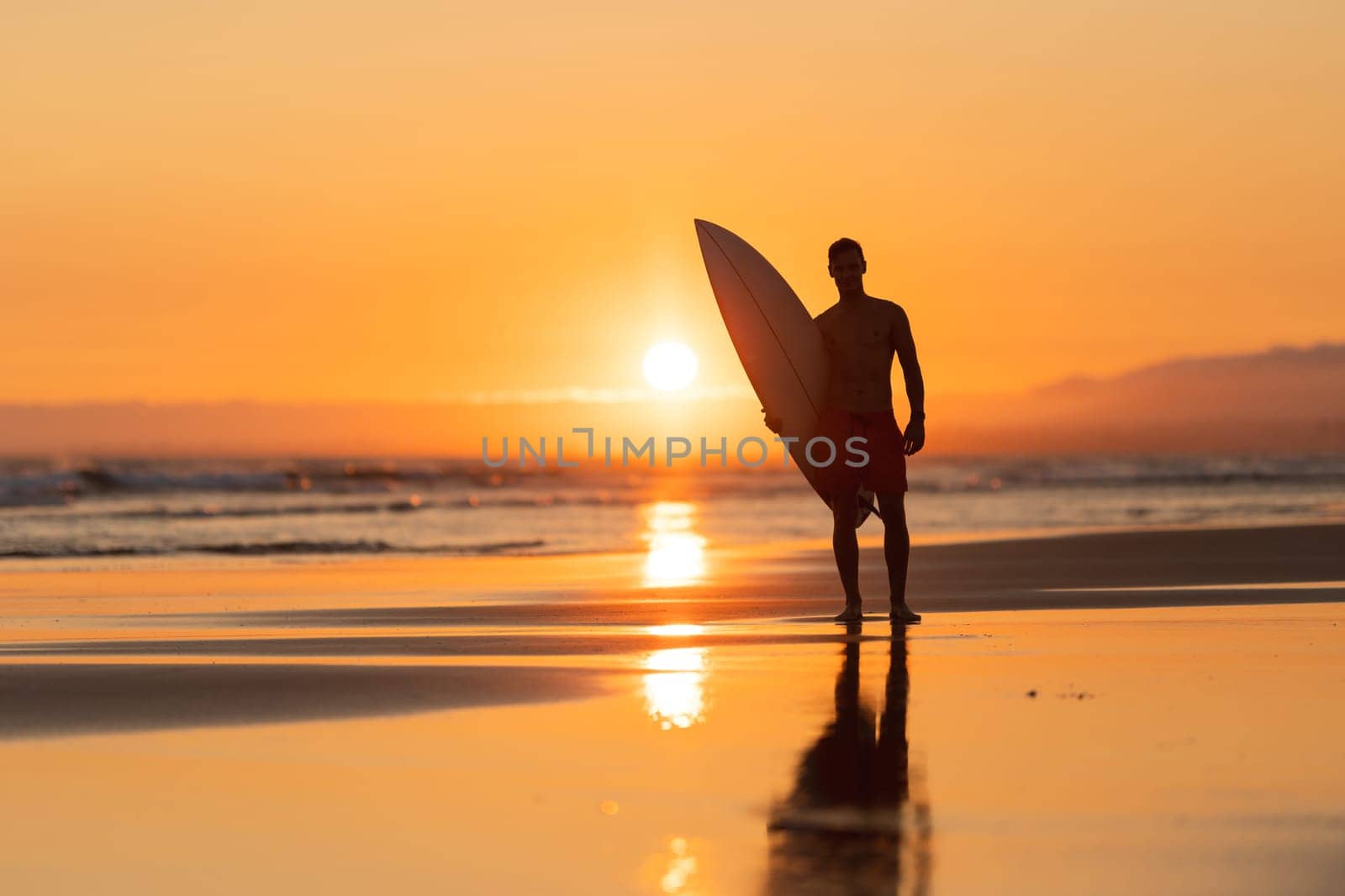Black silhouette of an attractive man on the shore holding a surfboard at orange sunset. Mid shot