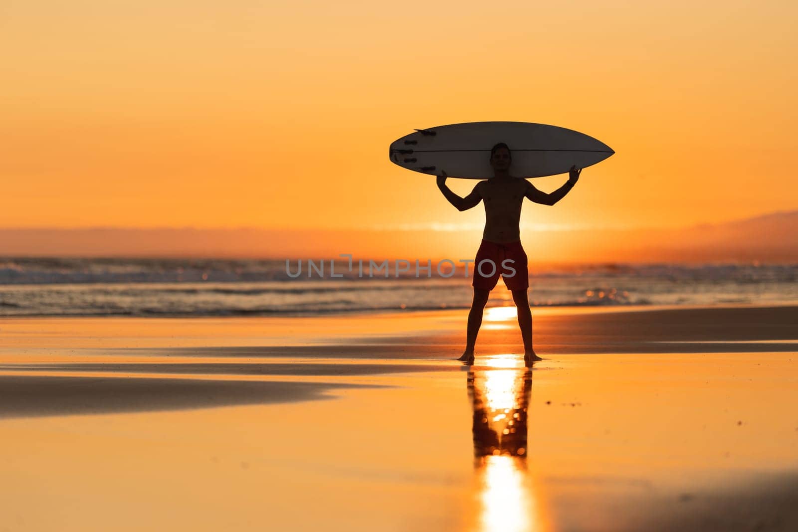 Black silhouette of an attractive man on the shore holding a surfboard above his shoulders at orange sunset by Studia72