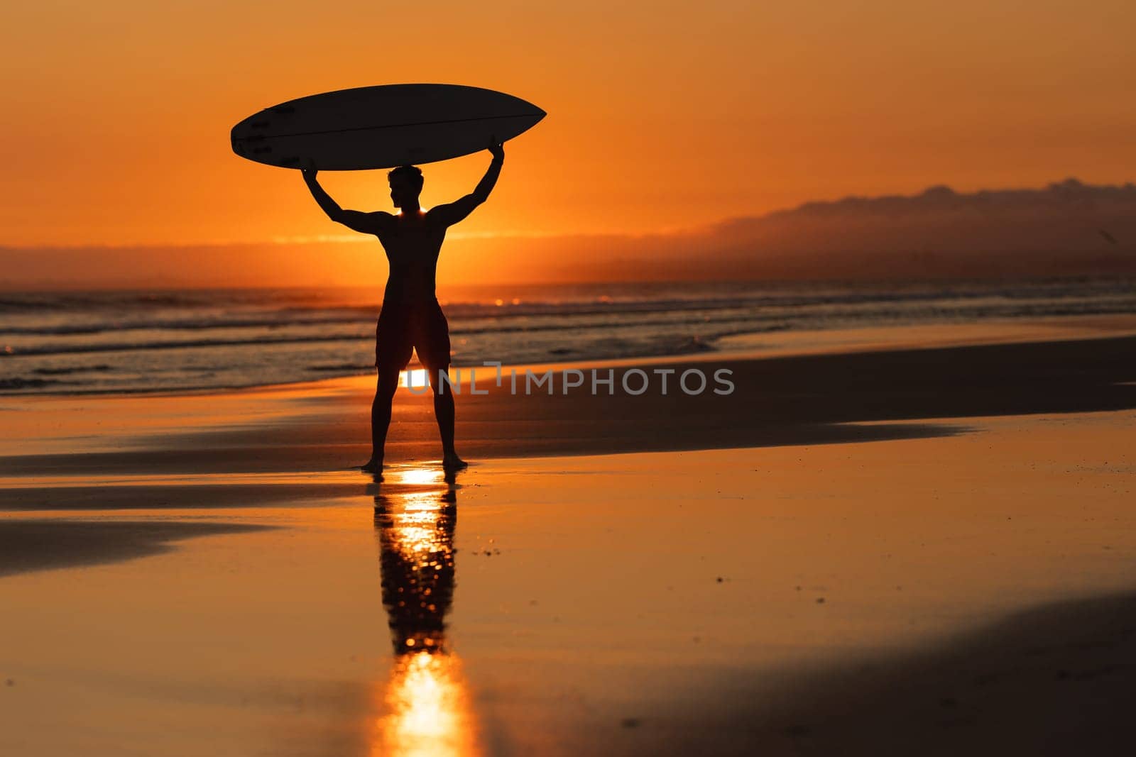 Silhouette of an athletic man standing on the shore holding a surfboard above his head at orange sunset by Studia72
