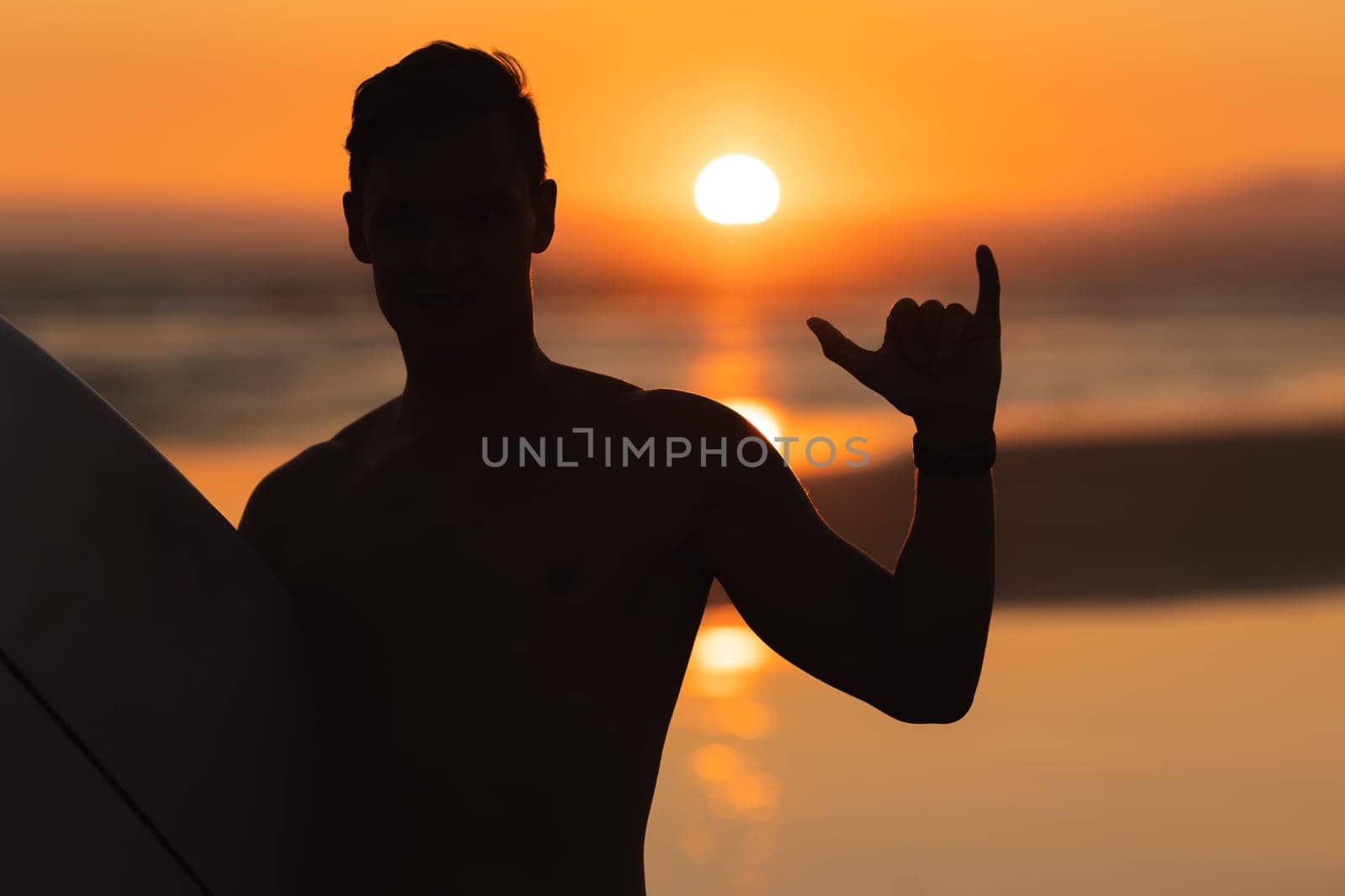 Black silhouette of an athletic man surfer showing shaka at sunset by Studia72