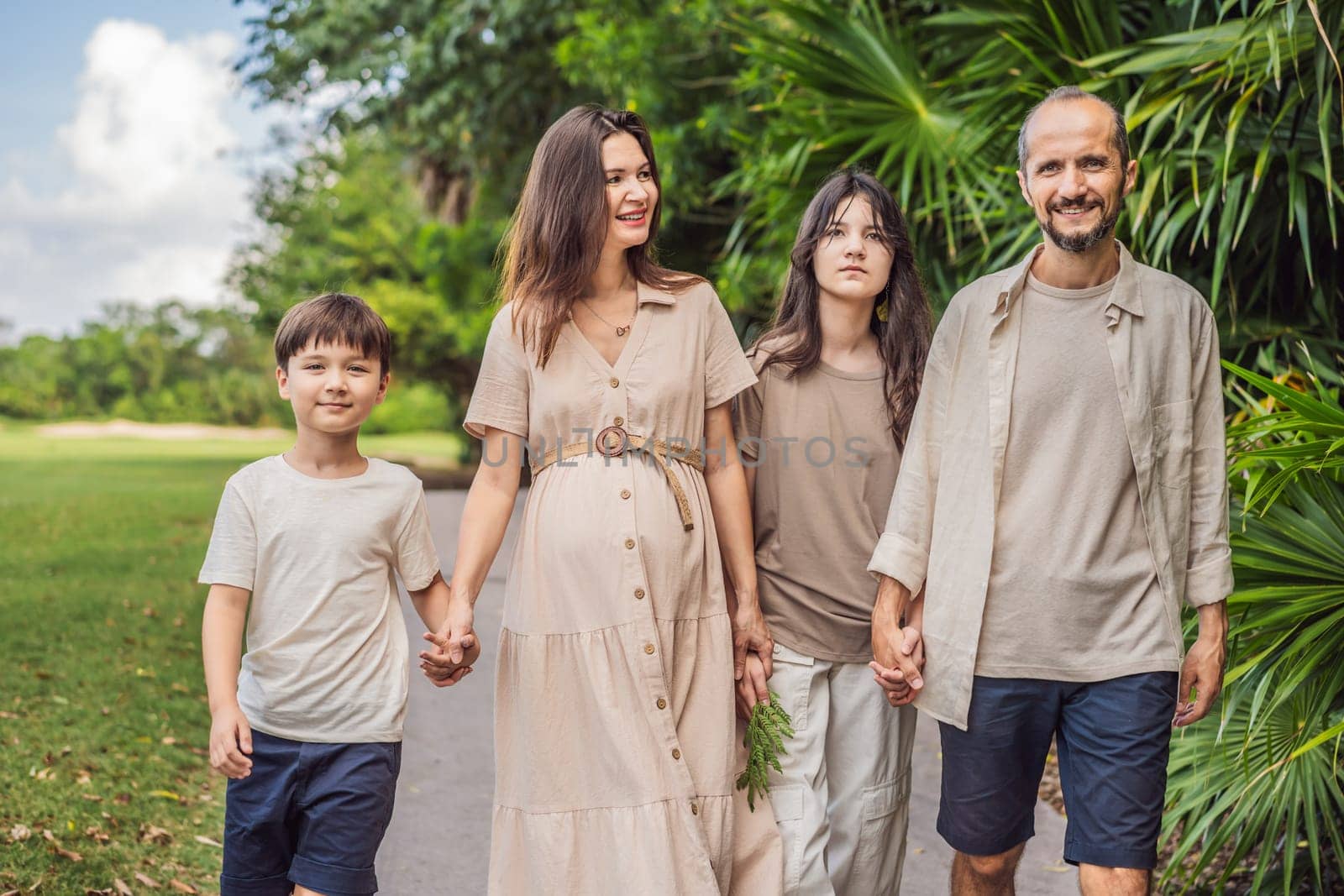 A loving family enjoying a leisurely walk in the park - a radiant pregnant woman after 40, embraced by her husband, and accompanied by their adult teenage children, savoring precious moments together amidst nature's beauty. Pregnancy after 40 concept by galitskaya