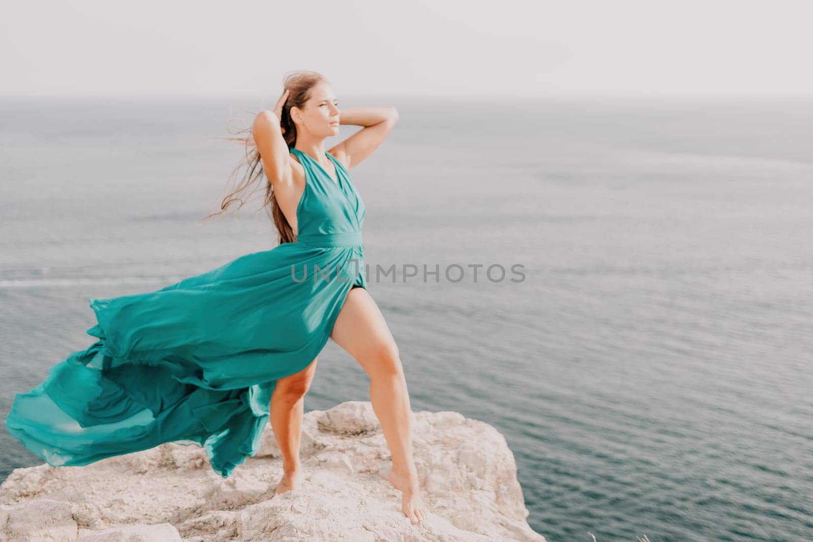 Woman travel portrait. Happy woman with long hair looking at camera and smiling. Close up portrait cute woman in a mint long dress posing on a volcanic rock high above the sea by panophotograph