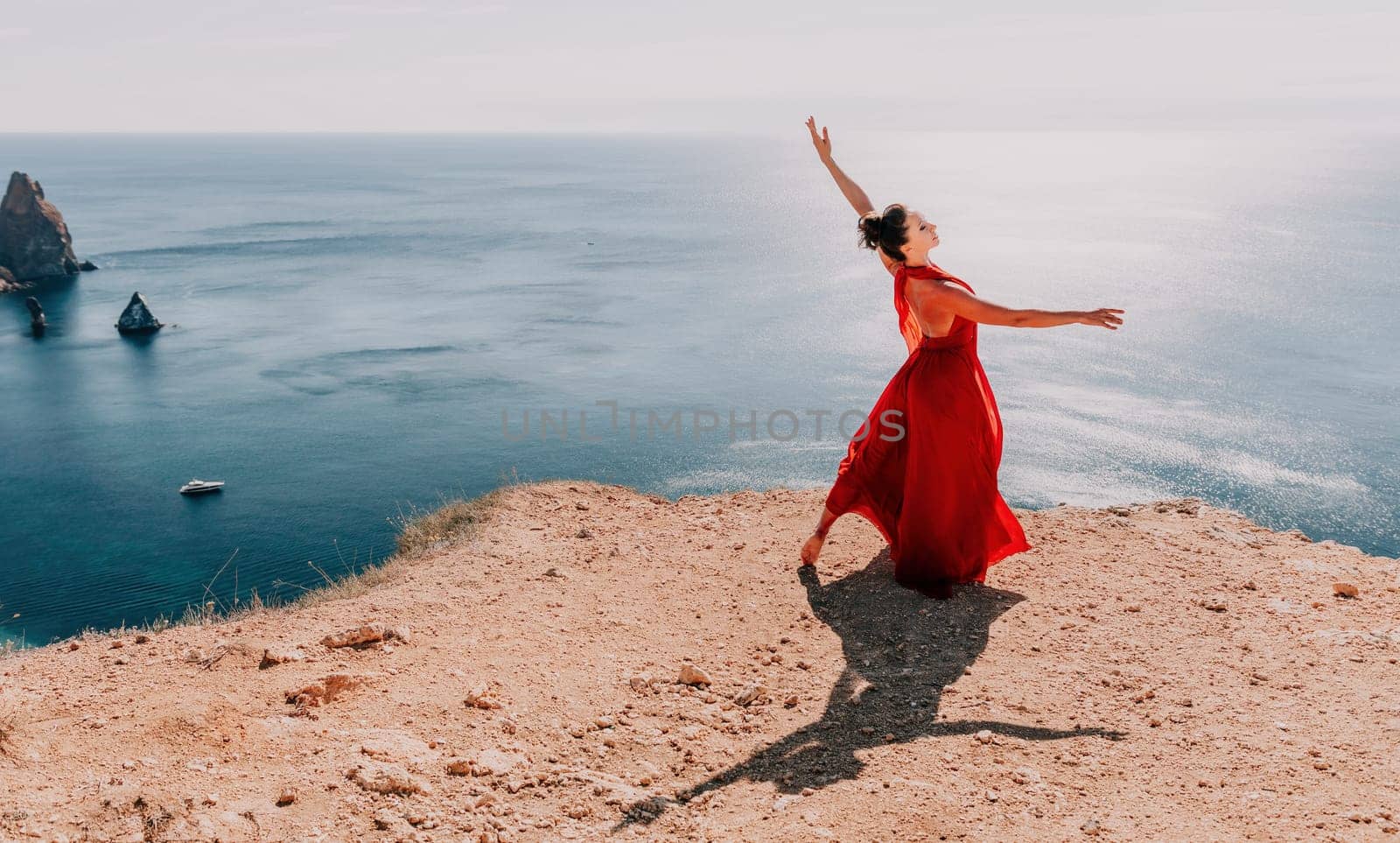 Side view a Young beautiful sensual woman in a red long dress posing on a rock high above the sea during sunrise. Girl on the nature on blue sky background. Fashion photo.