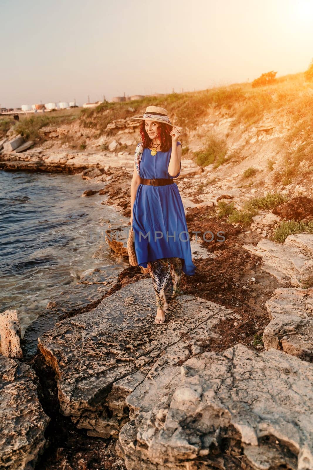 A woman in a dress, hat and with a straw bag is standing on the beach enjoying the sea. Happy summer holidays.