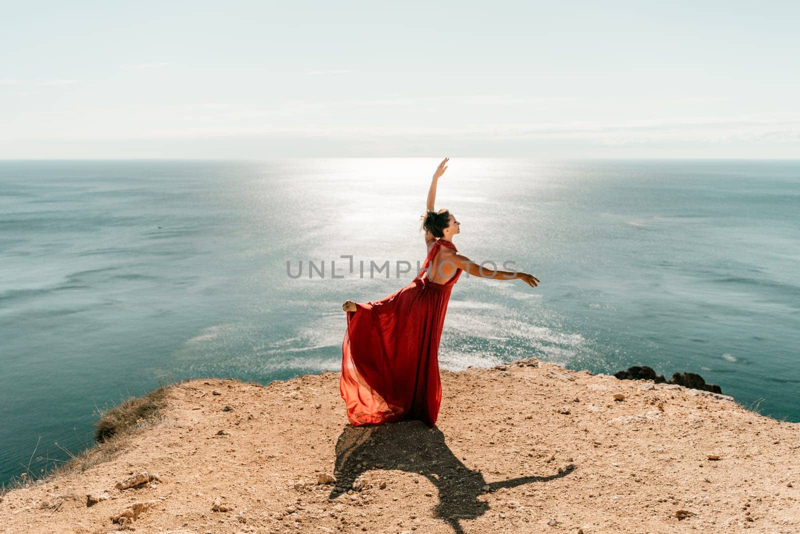 Woman red dress sea. Female dancer posing on a rocky outcrop high above the sea. Girl on the nature on blue sky background. Fashion photo