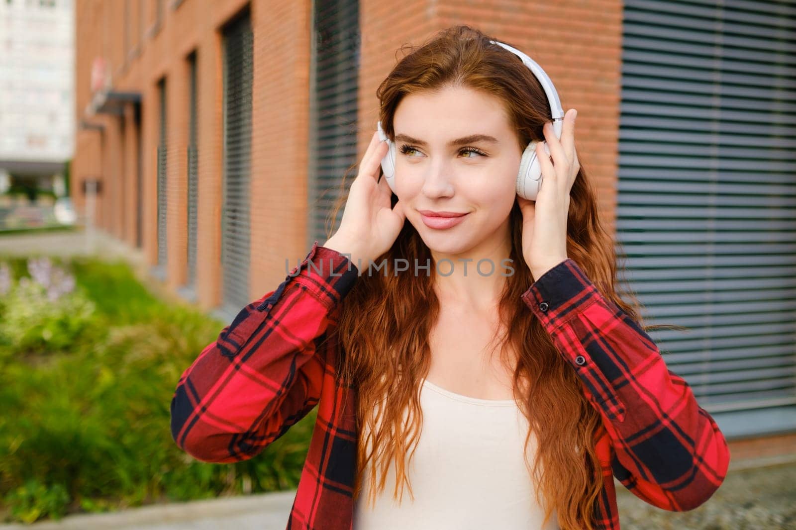 Portrait of smiling woman listening music via wireless headphones on the street, wearing checkered shirt.