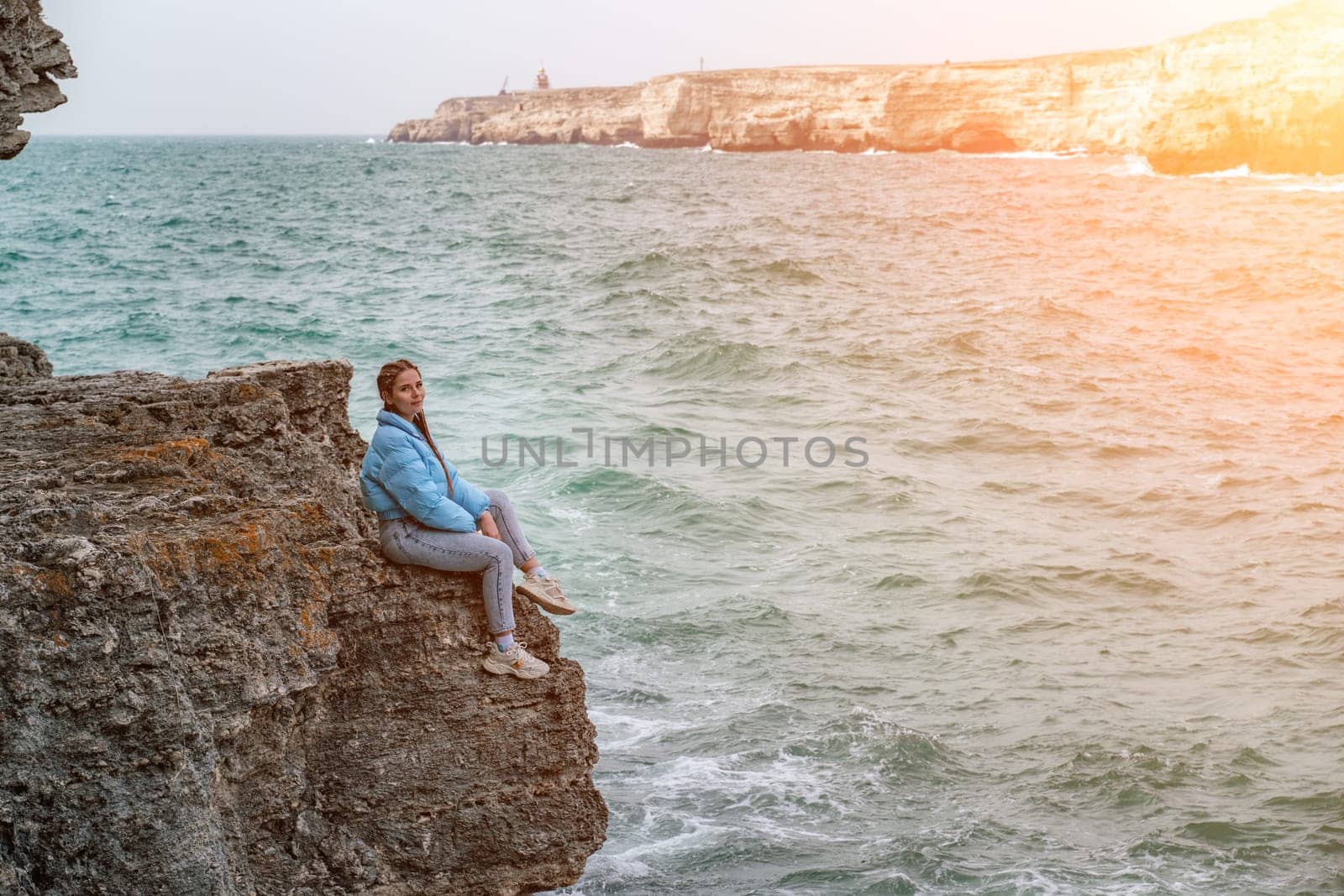 woman sea travel. A woman in a blue jacket sits on a rock above a cliff above the sea, looking at the stormy ocean. Girl traveler rests, thinks, dreams, enjoys nature by Matiunina