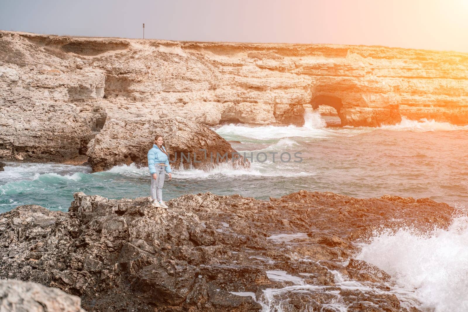 woman sea travel. A woman in a blue jacket stands on a rock above a cliff above the sea, looking at the stormy ocean. Girl traveler rests, thinks, dreams, enjoys nature by Matiunina