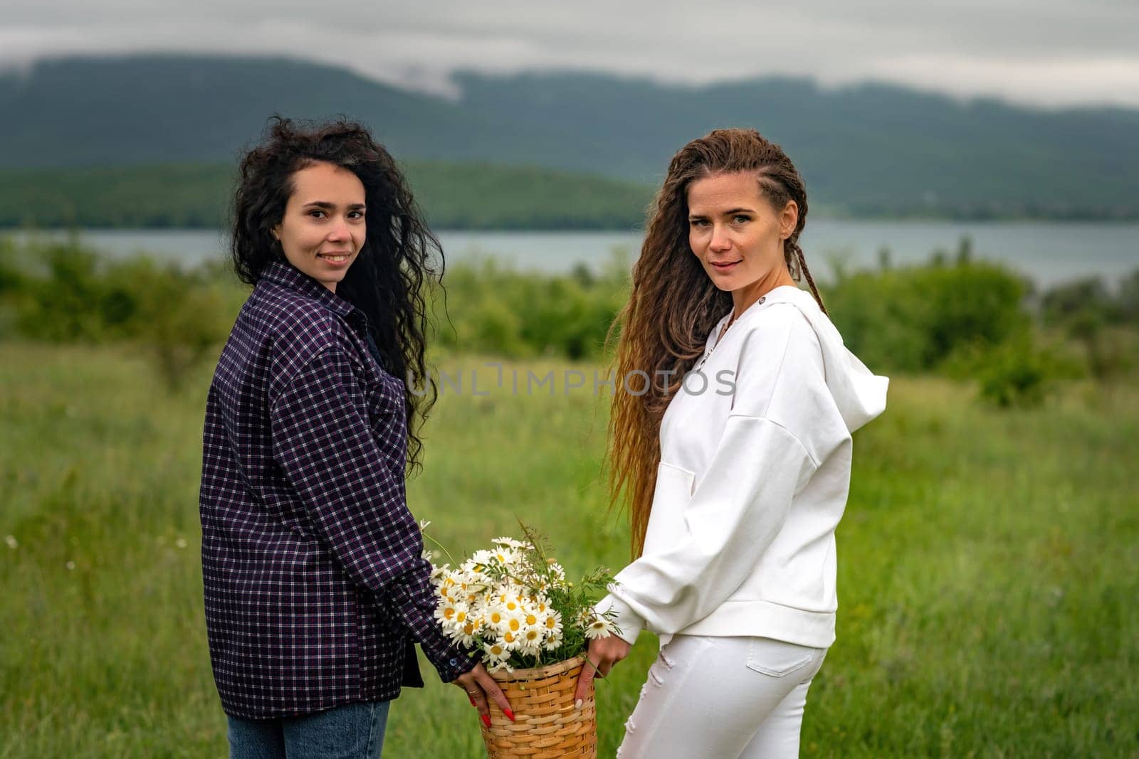 Two women enjoy nature in a field of daisies. Girlfriends hugging hold a bouquet of daisies and look at the camera. by Matiunina