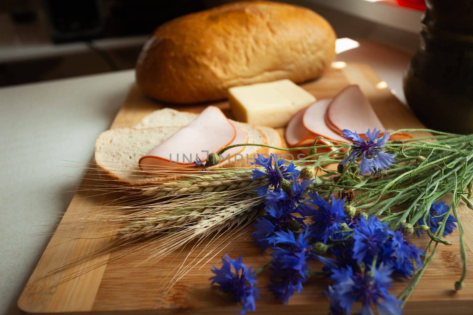 Ham sandwiches next to a loaf of bread with cereal and cornflower flowers lying on a wooden cutting board