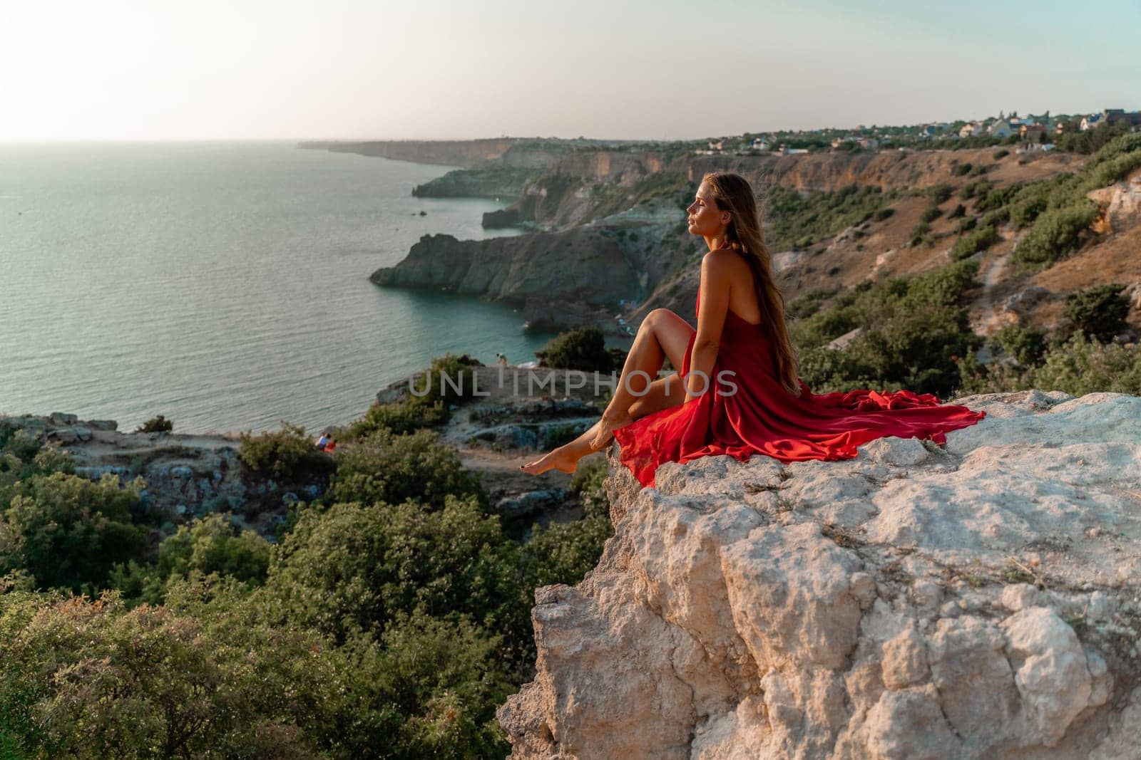 Woman sunset sea red dress, side view a happy beautiful sensual woman in a red long dress posing on a rock high above the sea on sunset. by Matiunina