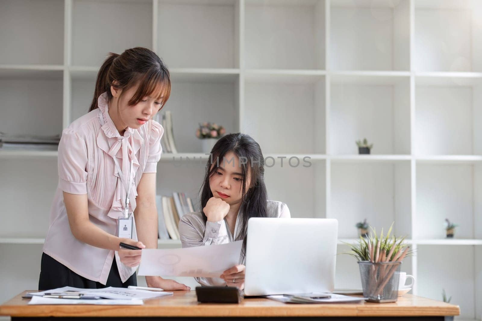 Two businesswomen colleagues working together look at laptop screen, talking about project, thinking, search solution, discuss business meet in modern office. Teamwork, workflow using tech by wichayada