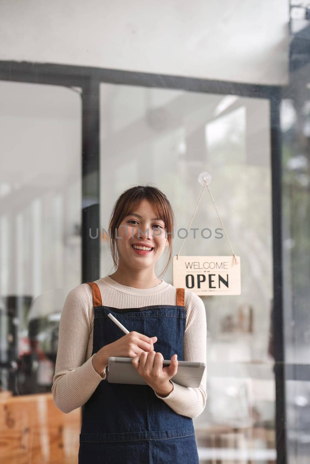 Successful small business owner. Beautiful girl with apron holding tablet standing in coffee shop restaurant. Portrait of asian woman barista cafe owner. by wichayada