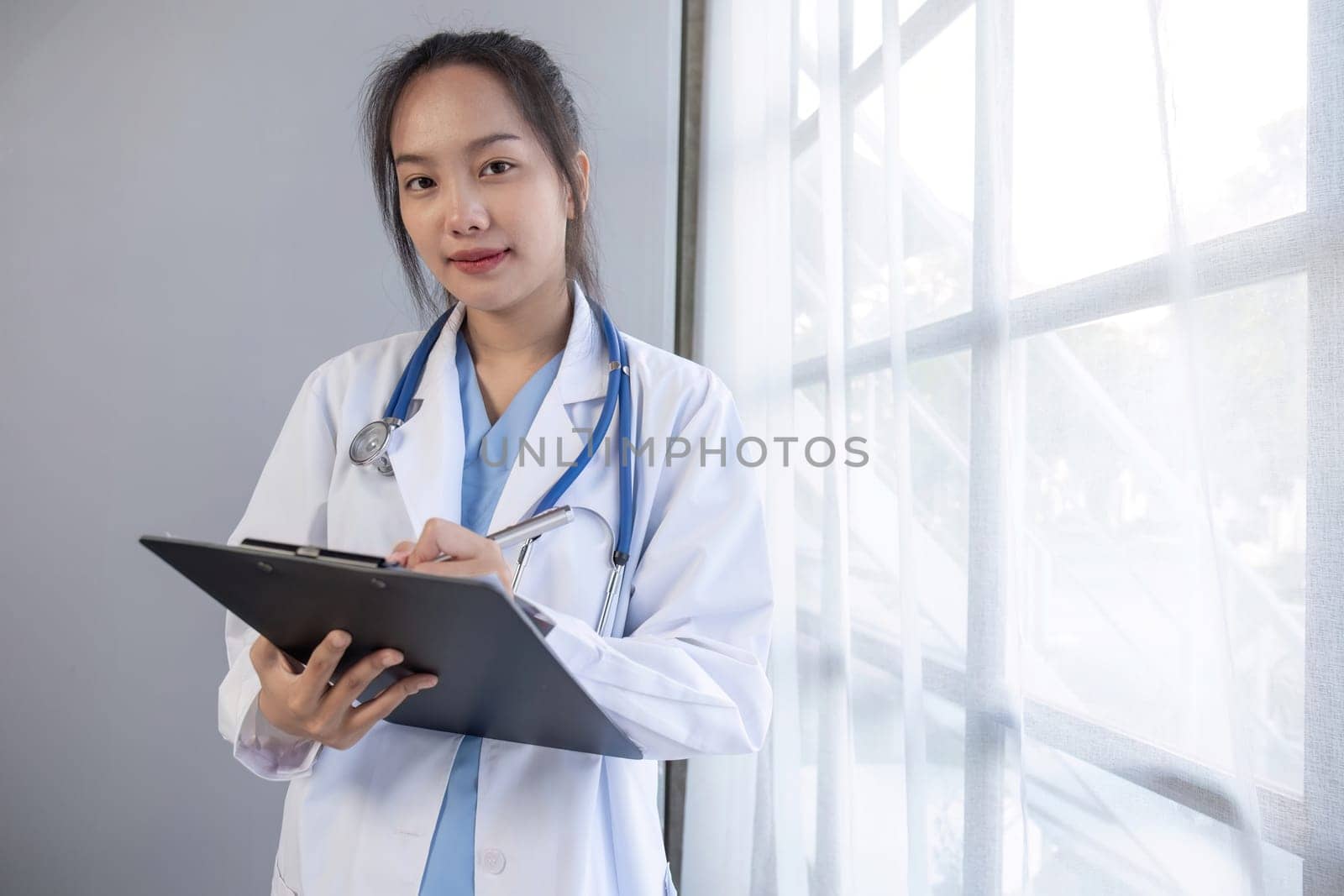 Portrait of female doctor wearing white coat with stethoscope holding clipboard and looking confidently to camera by wichayada