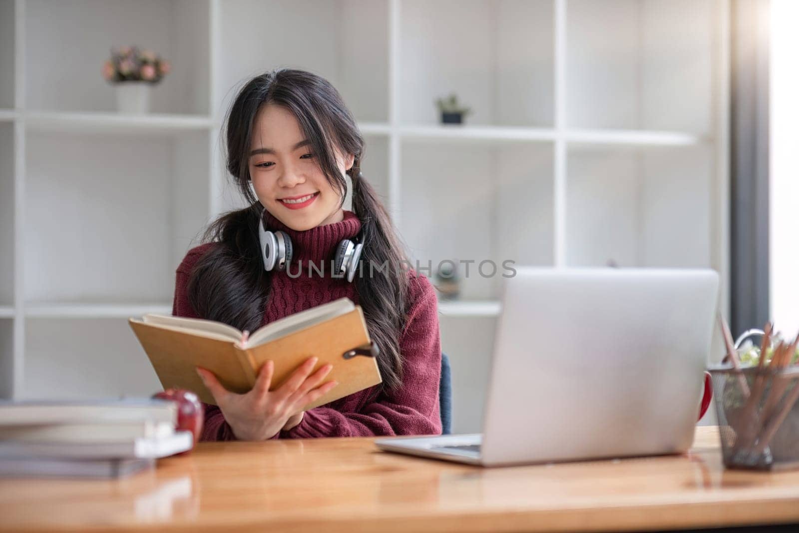 Asian female student studying online using headphones and laptop taking notes in notebook sitting at home table relaxing and changing places to school relax..