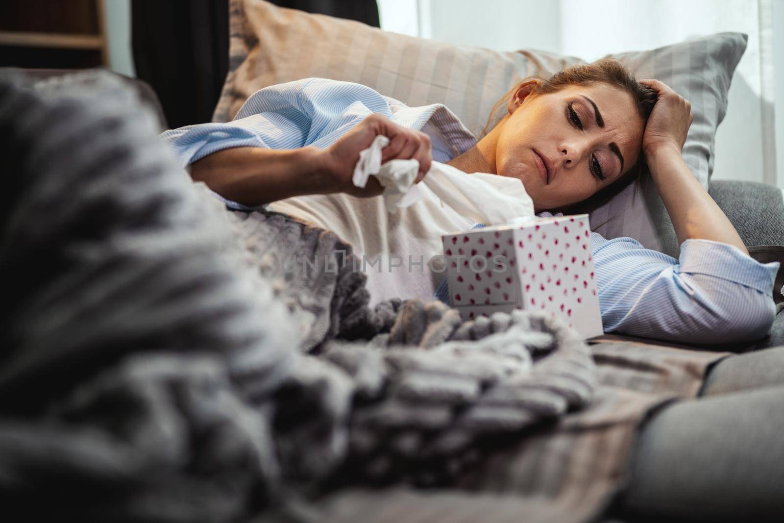 Young woman is lying sick at home couch and taking paper handkerchief for blowing nose.