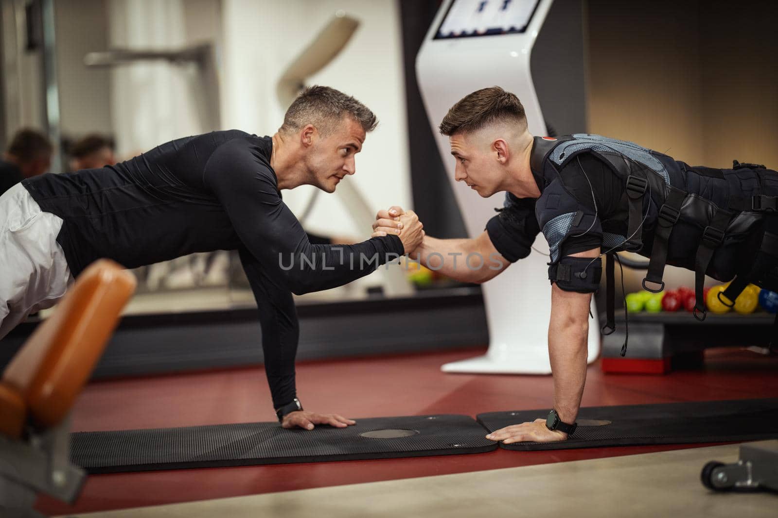 A young man is doing plank exercises with personal trainer in the gym, dressed in a black suit with an EMS electronic simulator to stimulate his muscles.