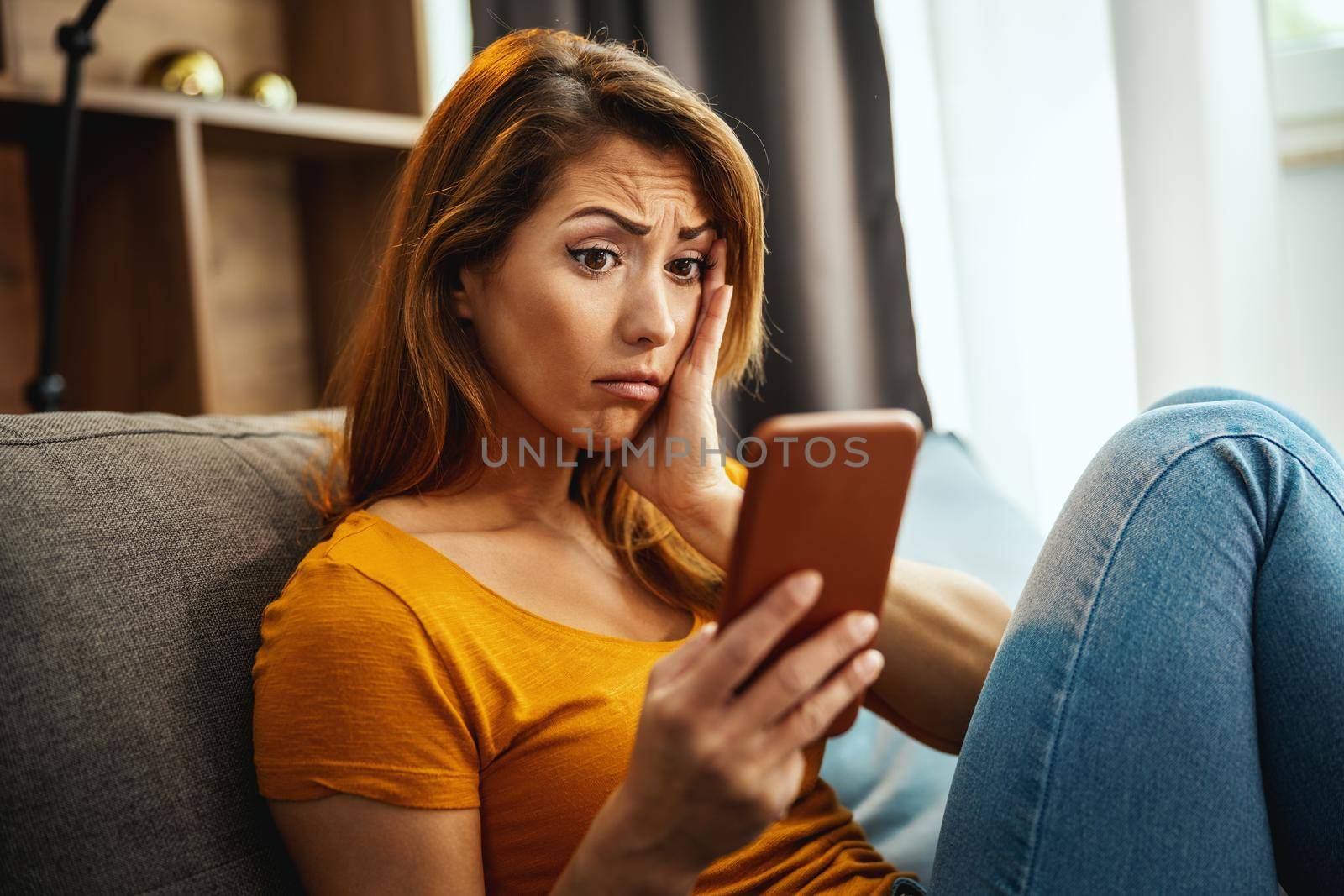 A worried young woman is sitting on the sofa and using smartphone to surfing social media at home.