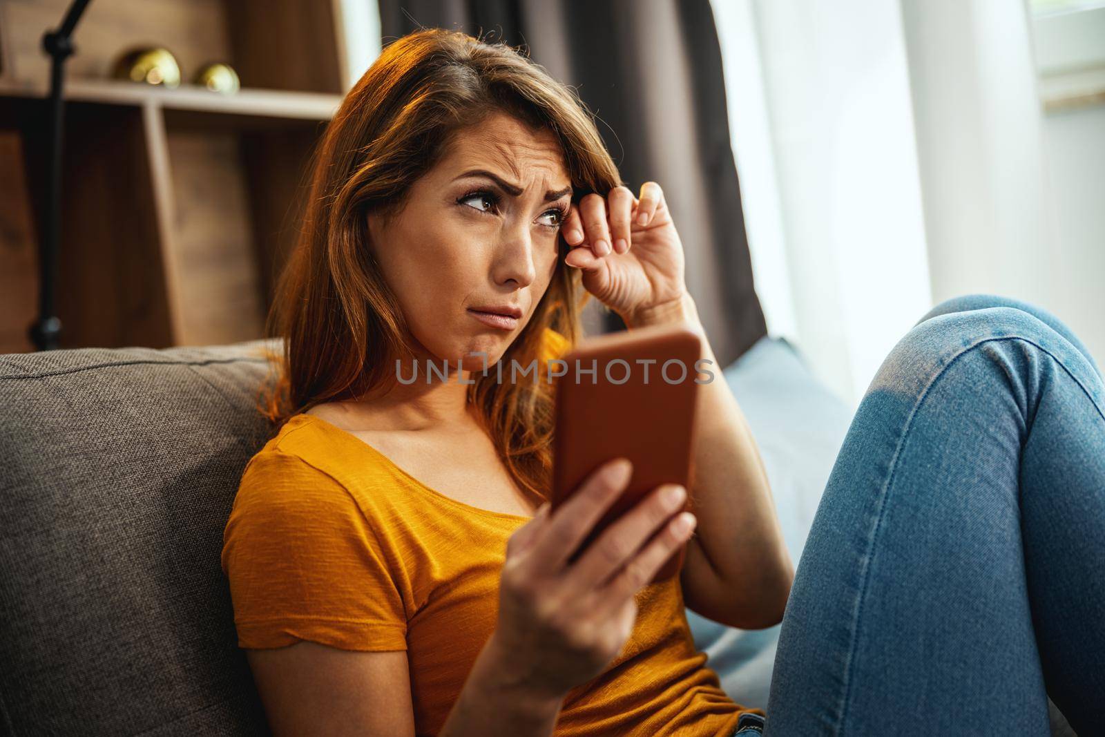 A unhappy young woman is sitting on the sofa and using smartphone to surfing social media at home.