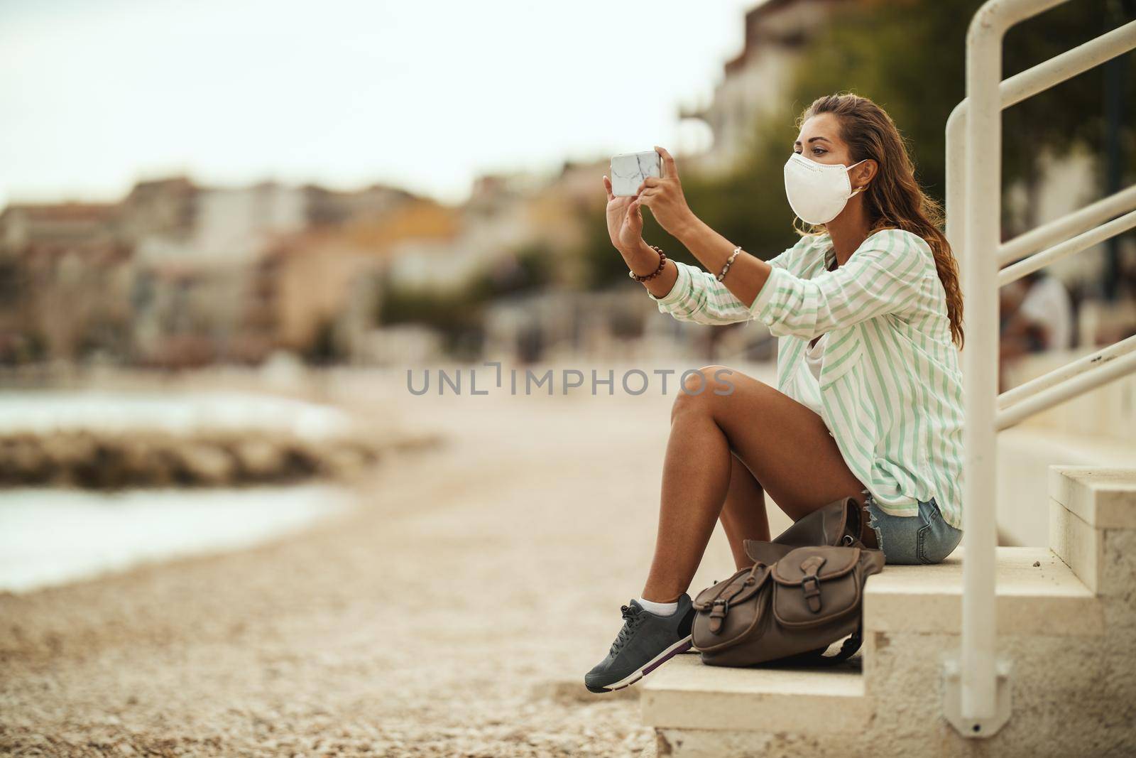 Shot of an attractive happy young woman wearing protective N95 mask and taking photo with a hers smartphone while enjoying a vacation on the beach during the COVID-19.