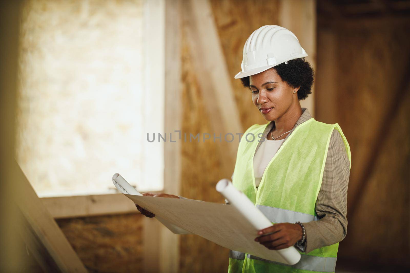 Shot of an African female architect checking blueprints at the construction site of a new wooden house. She is wearing protective workwear and white helmet.