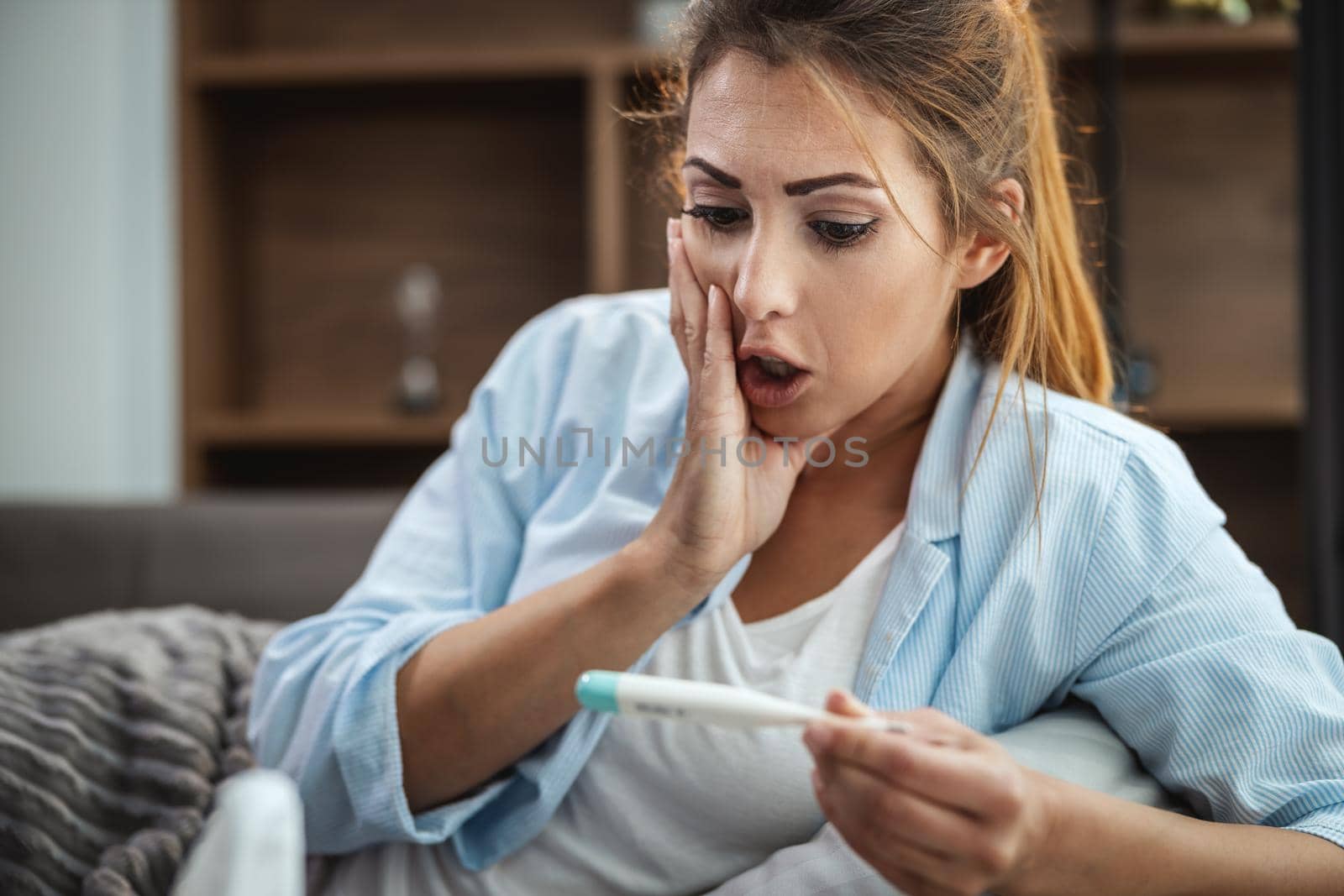 Young woman is lying sick at home couch and looking at thermometer.