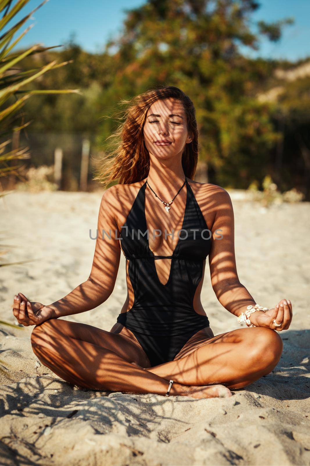 Attractive young woman is doing yoga and relaxing under the shade of a palm tree at the tropical beach.