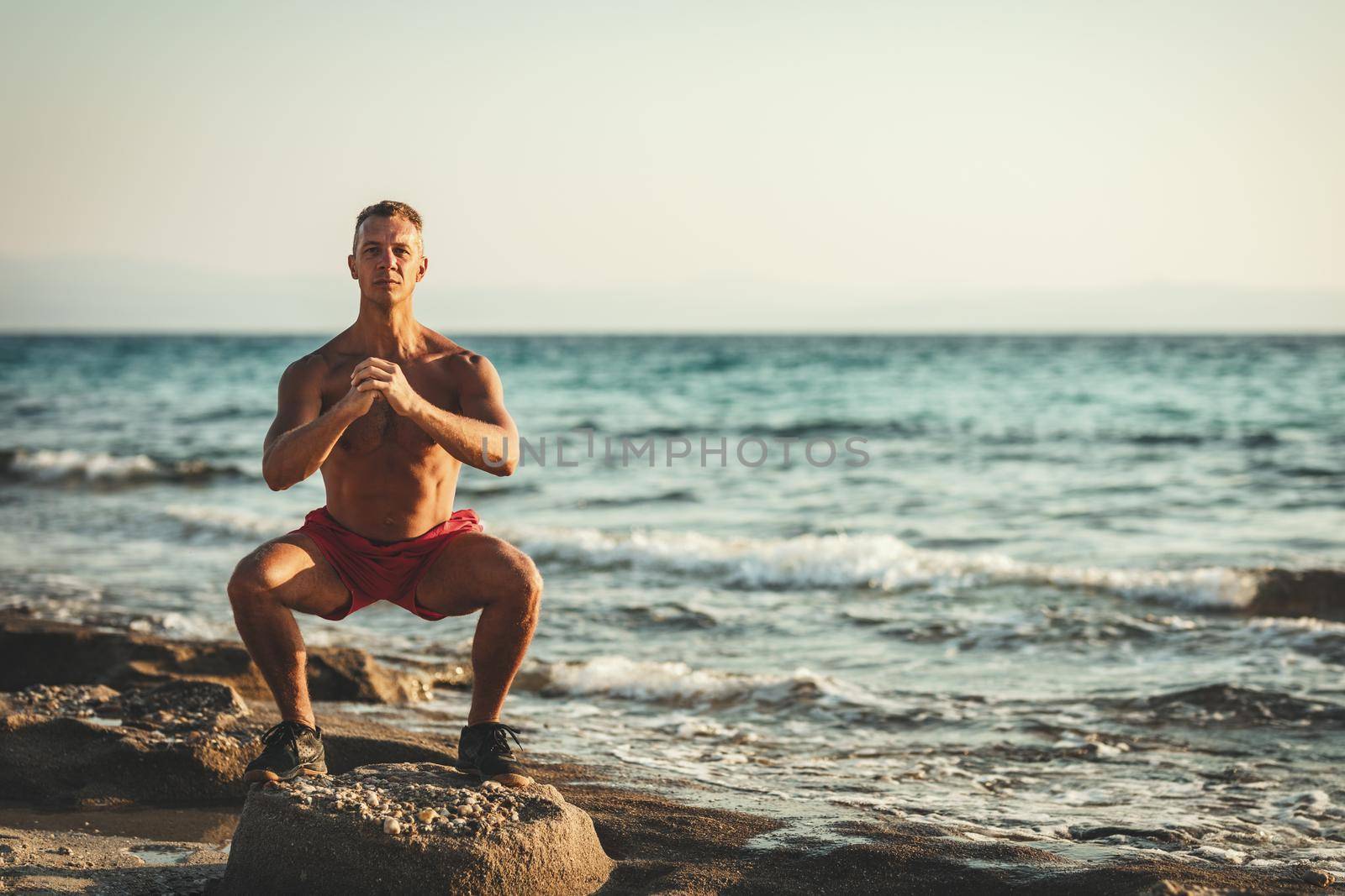 A handsome man is doing squat exercise at the sea beach in summer sunny day.
