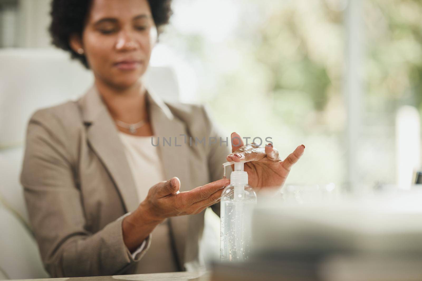 Cropped shot of an attractive African businesswoman sitting alone in her home office and using antiseptic gel to disinfect her hands during COVID-19 pandemic. Selective focus.