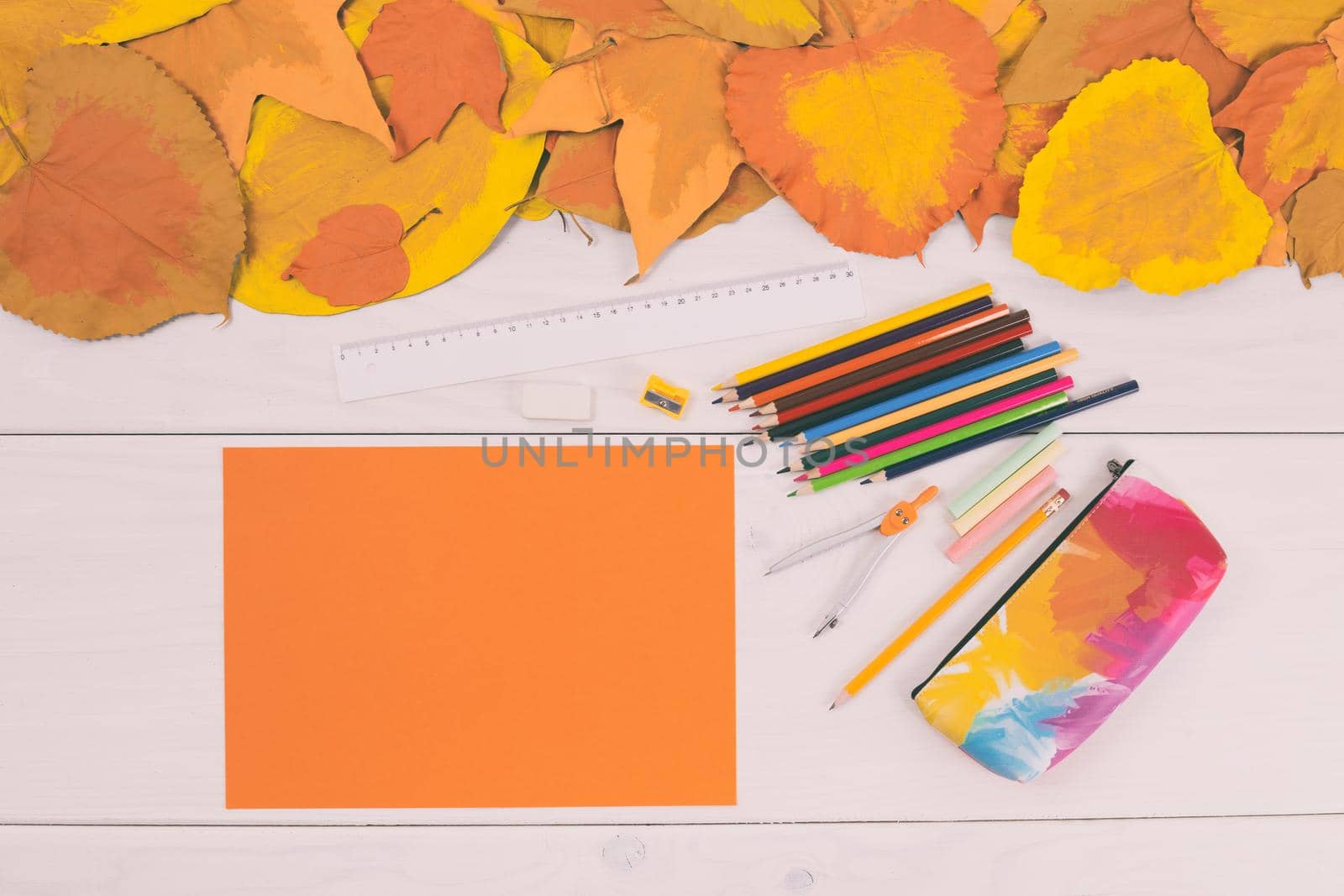 Empty orange paper and school supplies on wooden table with painted leaves.Toned image.