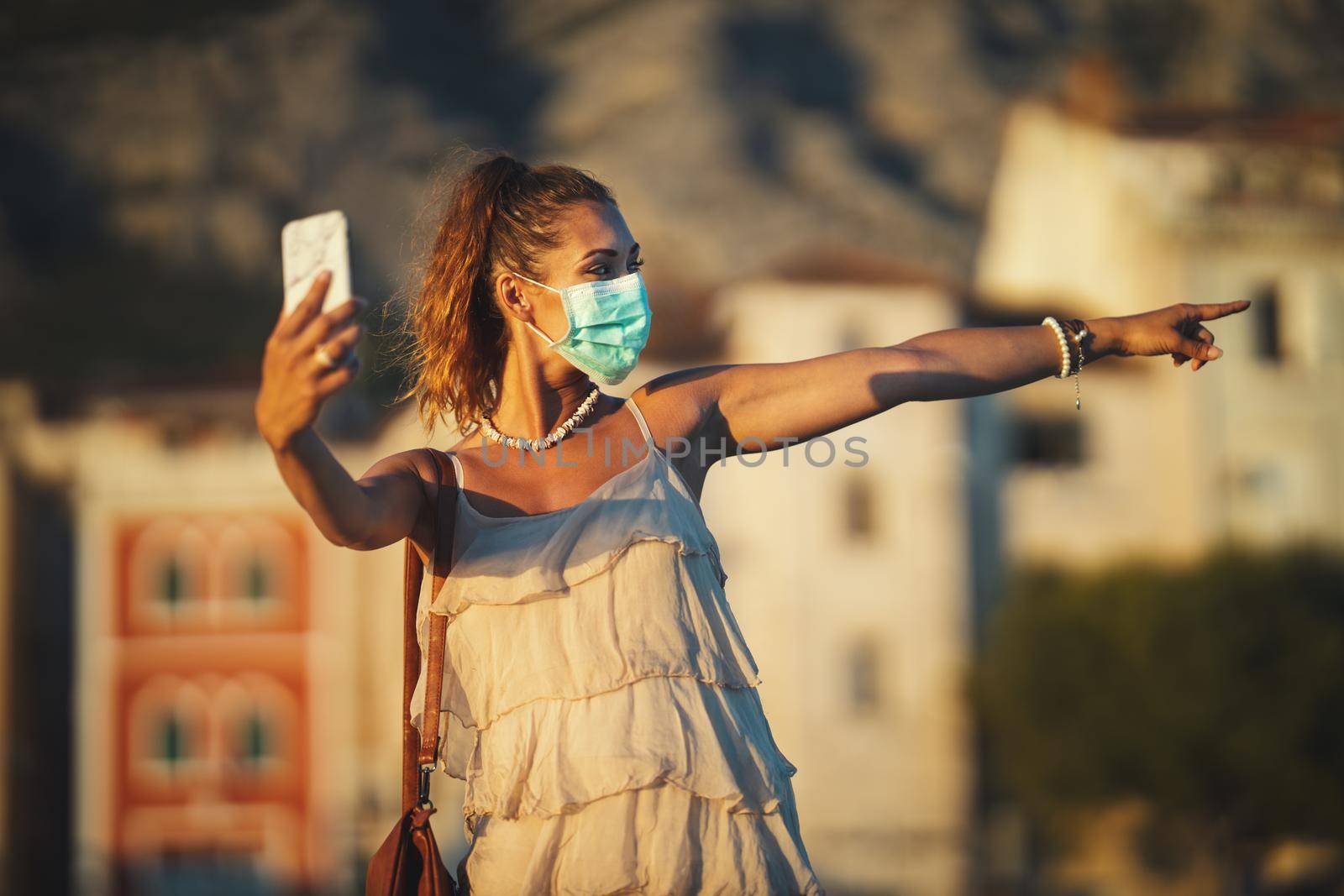 Shot of an attractive young woman wearing a surgical mask and making video call with her smartphone while exploring the wonderful city of Meditteranian.