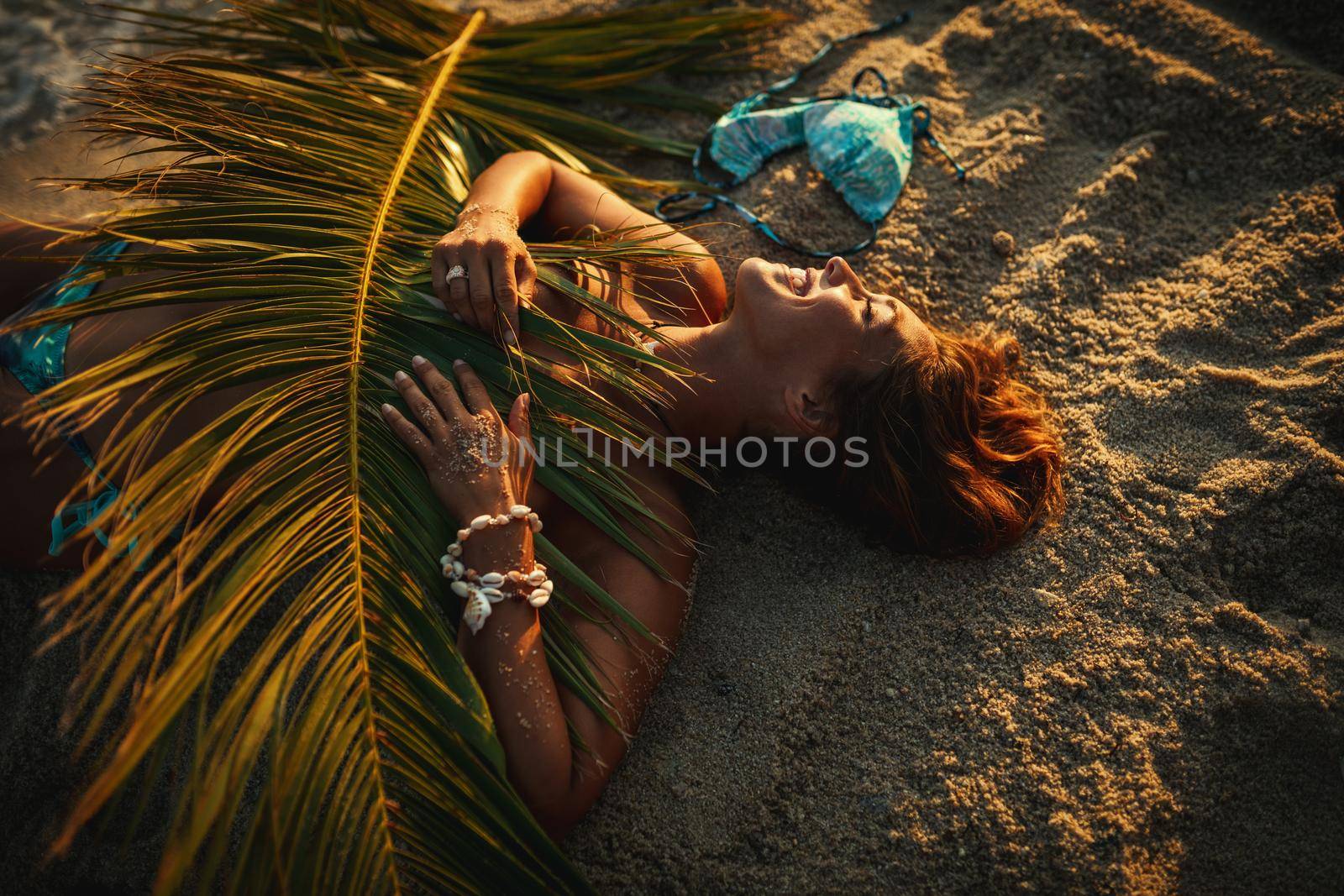 Shot of an attractive young woman is lying on the tropical sandy beach covered with palm tree leaf.