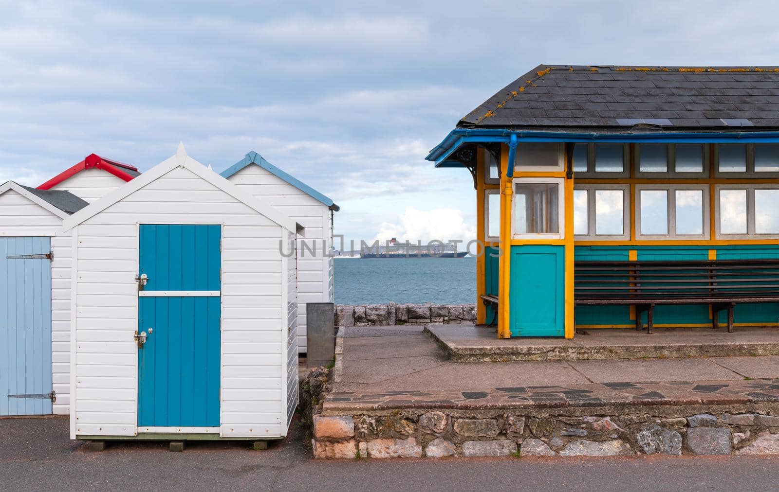 Colorful small beach houses. Multicolored beach sheds. Variety of painted beach shacks. Cruise ship on water in background. Beach huts. Torbay, South Devon. UK. by Qba