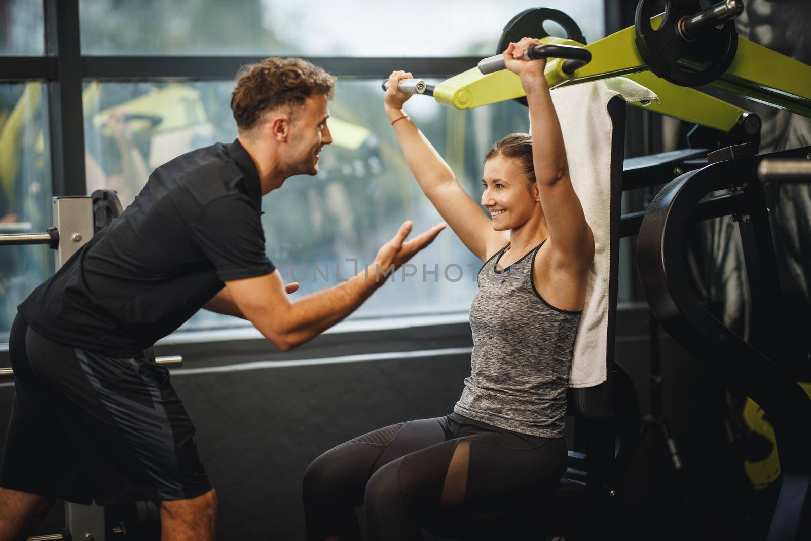 Shot of a muscular young woman in sportswear working out with personal trainer at the gym machine. She is pumping up her shoulder muscule with heavy weight.