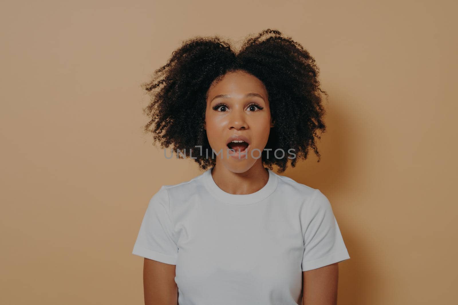 Photo of amazed stunned dark skinned female looking at camera with opened mouth, shocked african woman in white tshirt with surprised face expression posing against beige background