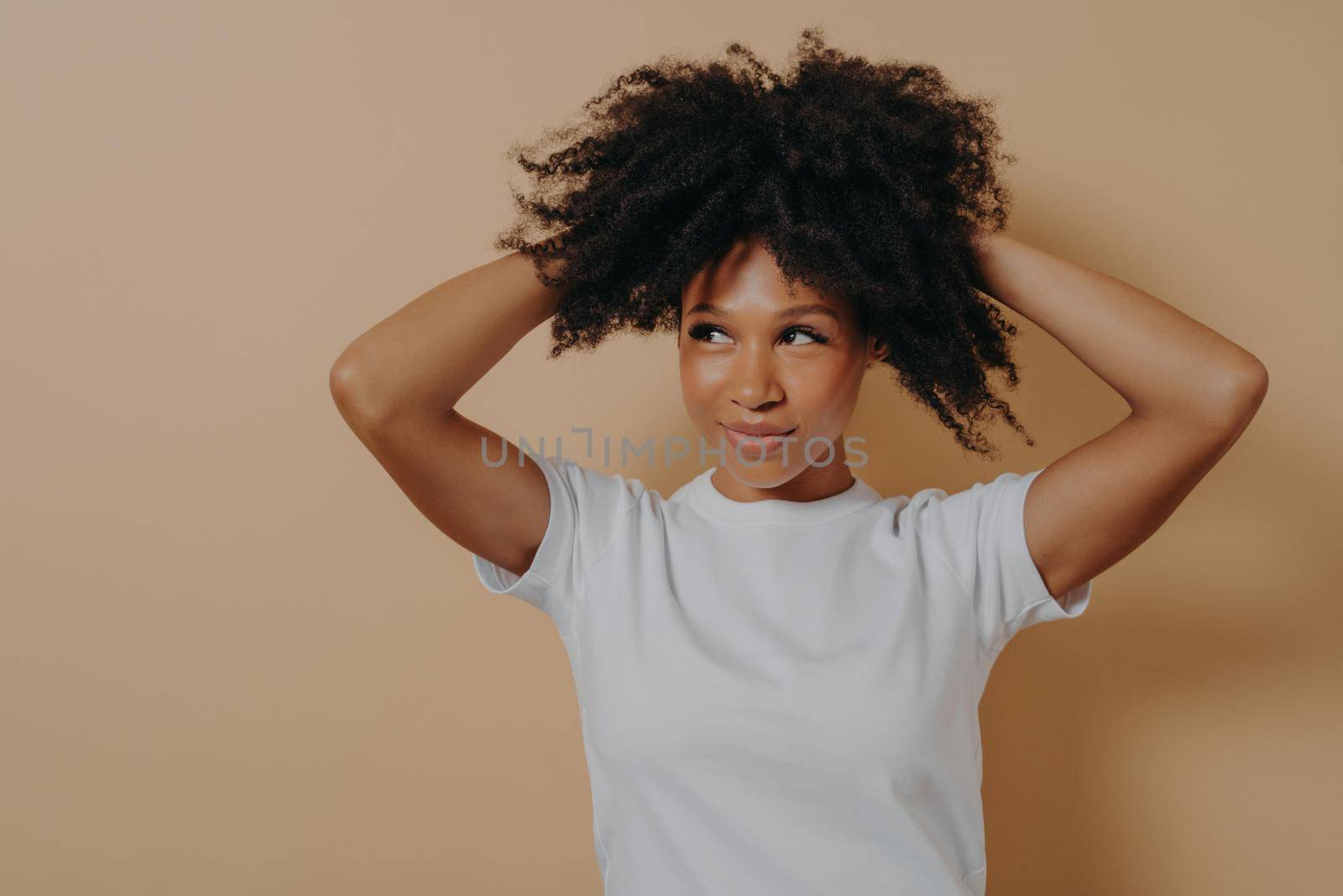 Young attractive sensual african woman touching with hands curly hair, isolated on beige background by vkstock