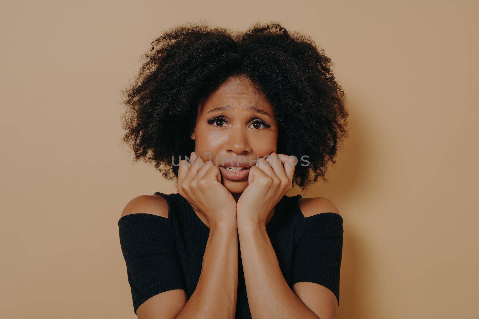 Scared mixed race female with curly hairstyle looking nervously and feeling stress, african woman being afraid of something while posing against beige background. Fear and anxiety concept