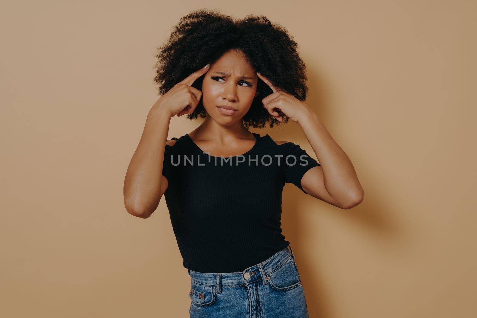 Young thoughtful mixed race woman with curly hair holds fingers on temples and trying to concentrate, demonstrating thinking process while posing isolated over beige wall. Concentration concept