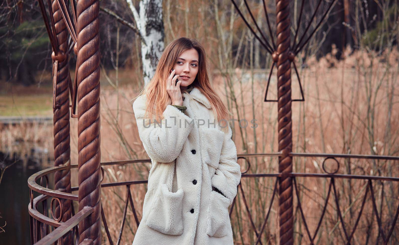 Happy beautiful young woman talking on mobile phone in the gazebo on the background of the pond