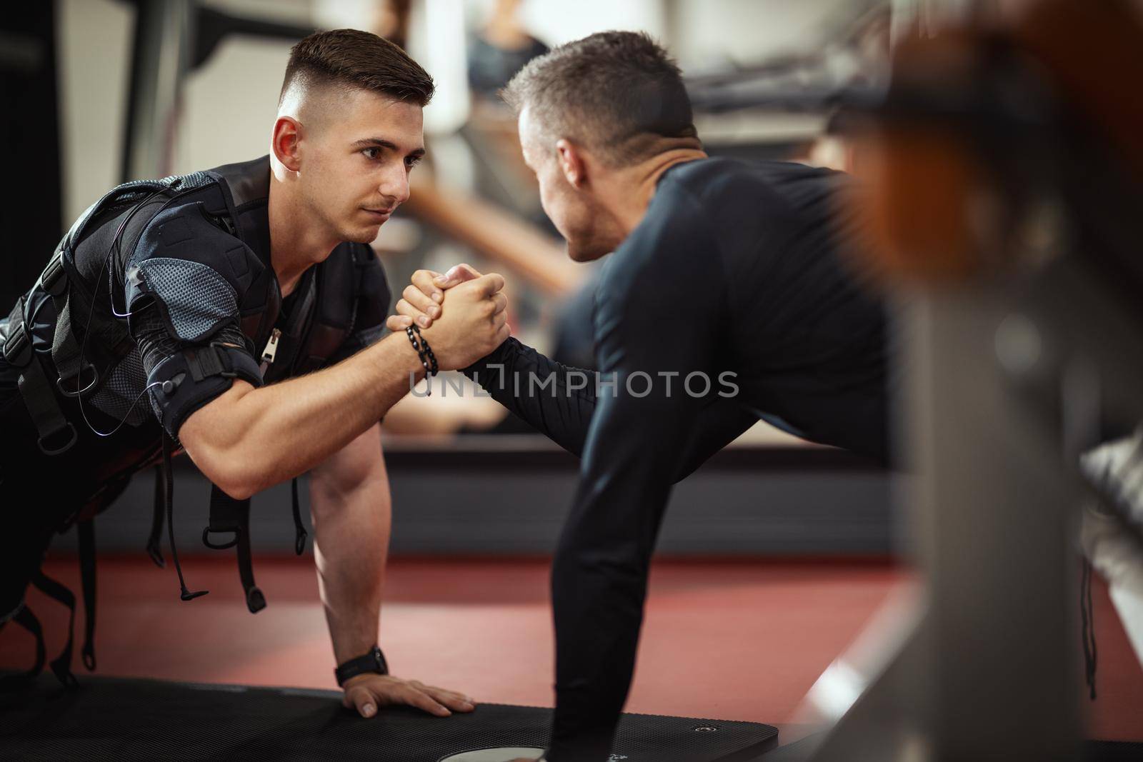 A young man is doing plank exercises with personal trainer in the gym, dressed in a black suit with an EMS electronic simulator to stimulate his muscles.