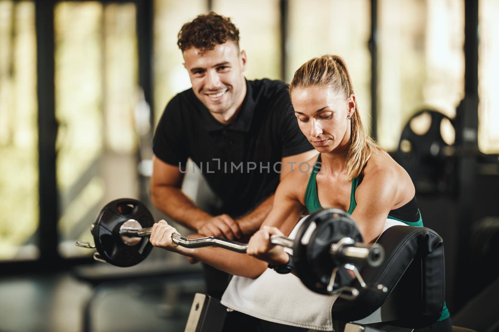 Shot of a muscular young woman in sportswear working out with personal trainer at the gym. She is pumping up her biceps muscule with heavy weight.